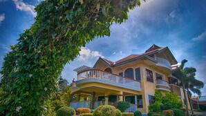 Une grande maison à deux étages, dotée d’un balcon et entourée de jardins et d’arbres bien entretenus.