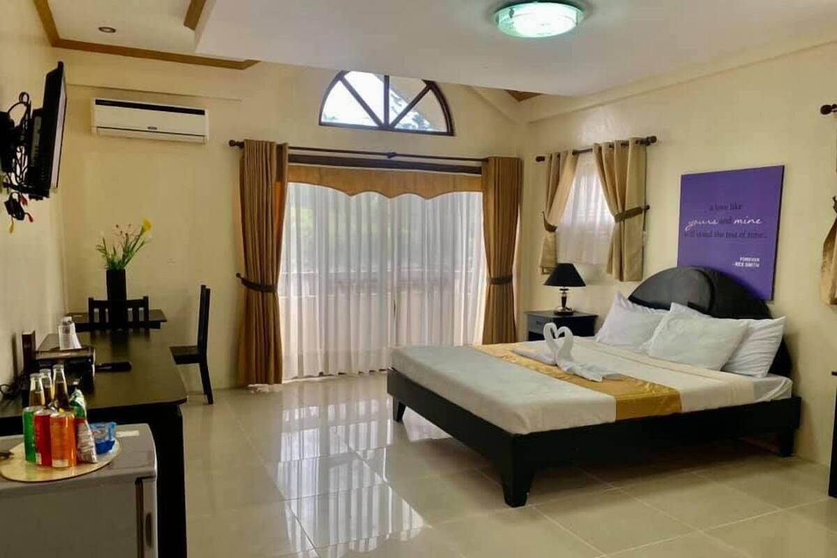 A hotel room with a bed, a desk with a chair, a small table with bottles, and a window with curtains.