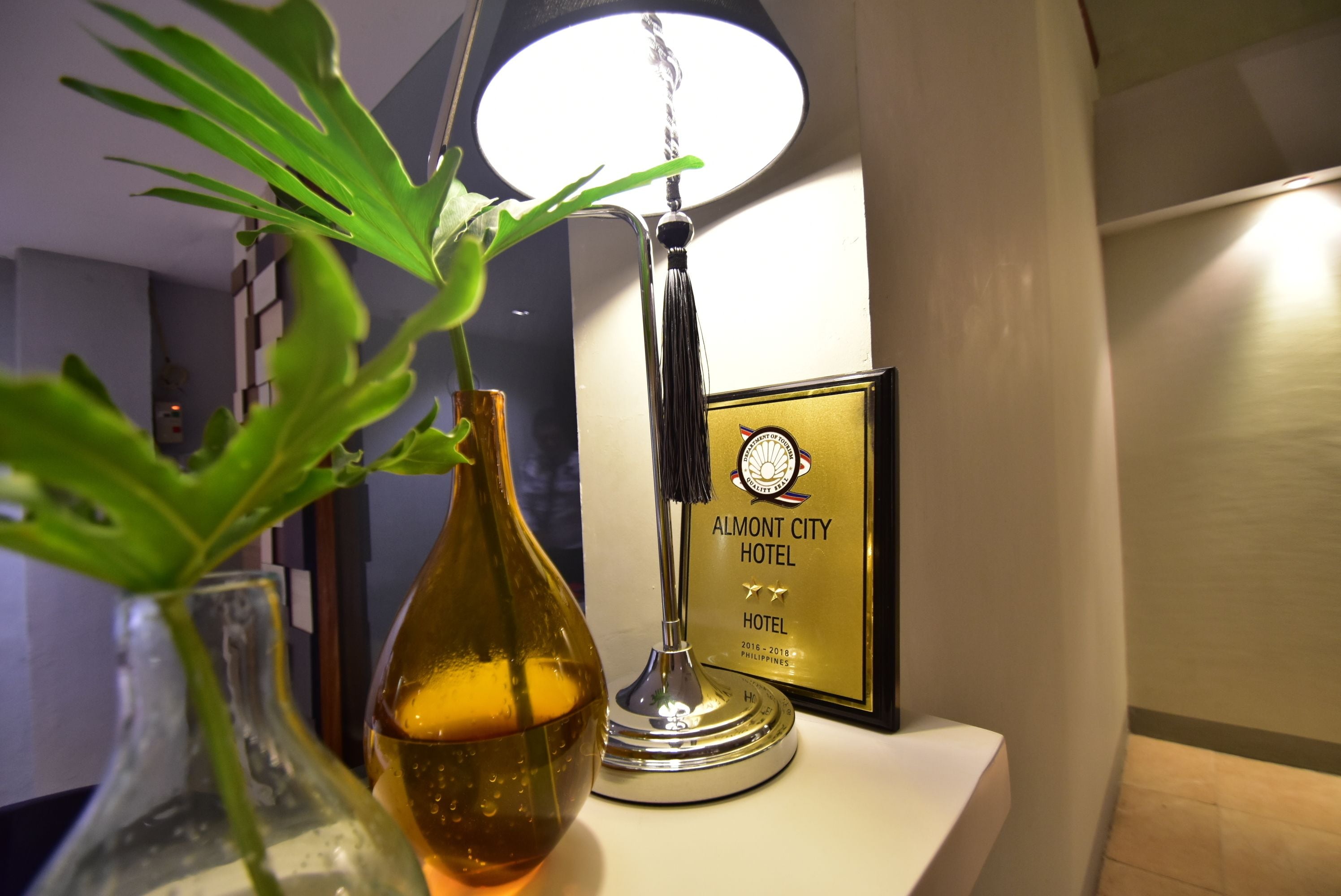 Eine gerahmte Auszeichnung für das Almont City Hotel, eine transparente Glasvase mit Pflanze und eine Lampe auf einer weißen Oberfläche.