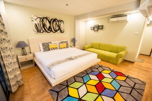 Una habitación de hotel con una cama, un sofá verde, un tapete colorido, una obra de arte en la pared y un equipo de aire acondicionado.