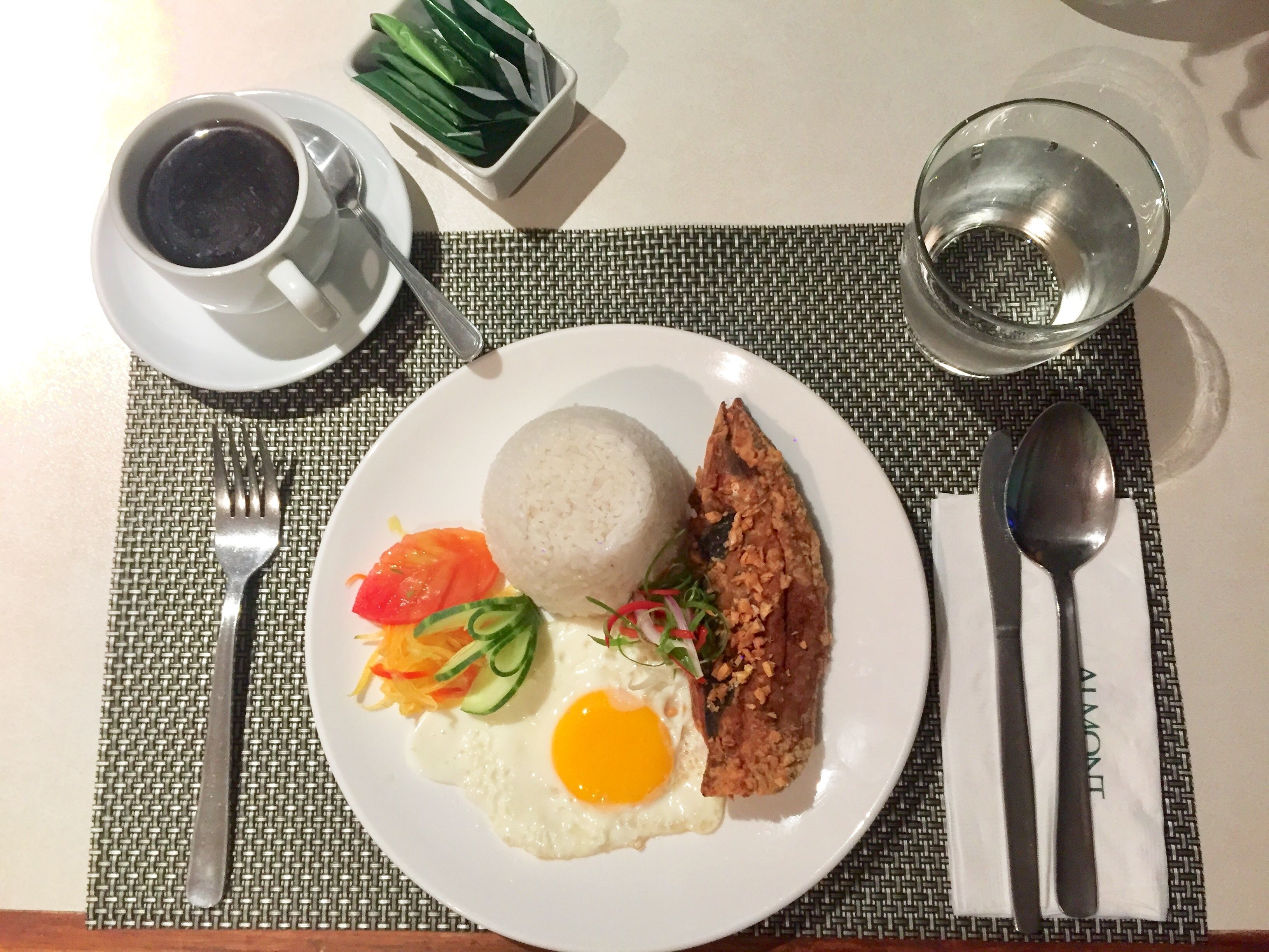 Ein Teller mit Reis, Spiegelei, gebratenem Fleisch und Gemüse, eine Tasse Kaffee und ein Glas Wasser auf einem Tisch.