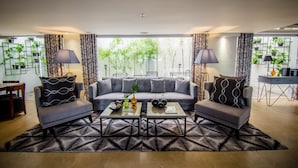 A modern living room with a grey sofa, two armchairs, a coffee table, and a patterned rug. There are two lamps, a bookshelf with plants, and a view of trees through large windows. - Almont City Hotel (Butuan)