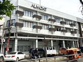 A multi-story building with the sign "ALMONT CITY HOTEL BUUTAN" indicating it is a hotel. There are several vehicles parked in front, including a white van and a motorcycle.