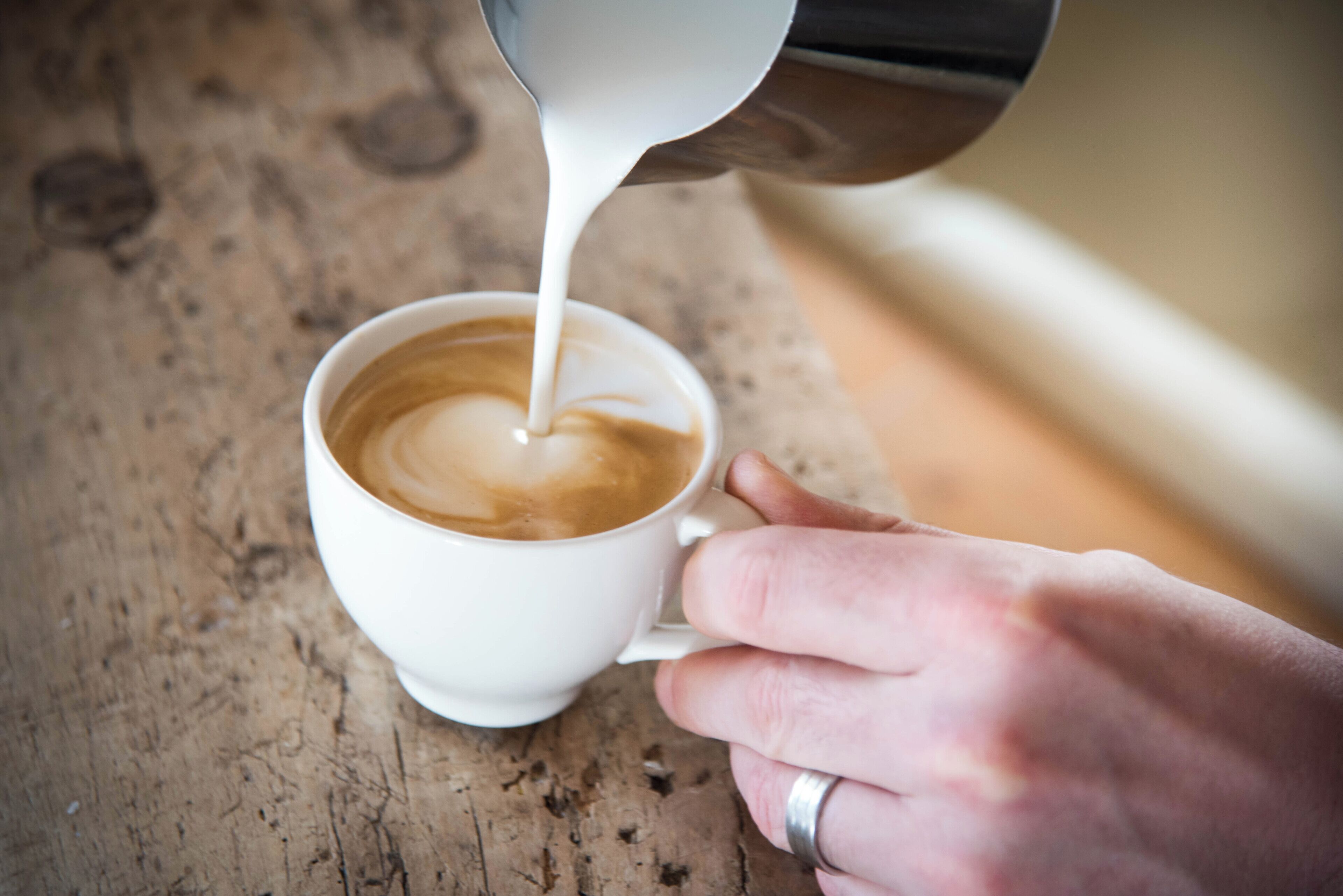 A hand holding a white cup, pouring milk into a cup of coffee.