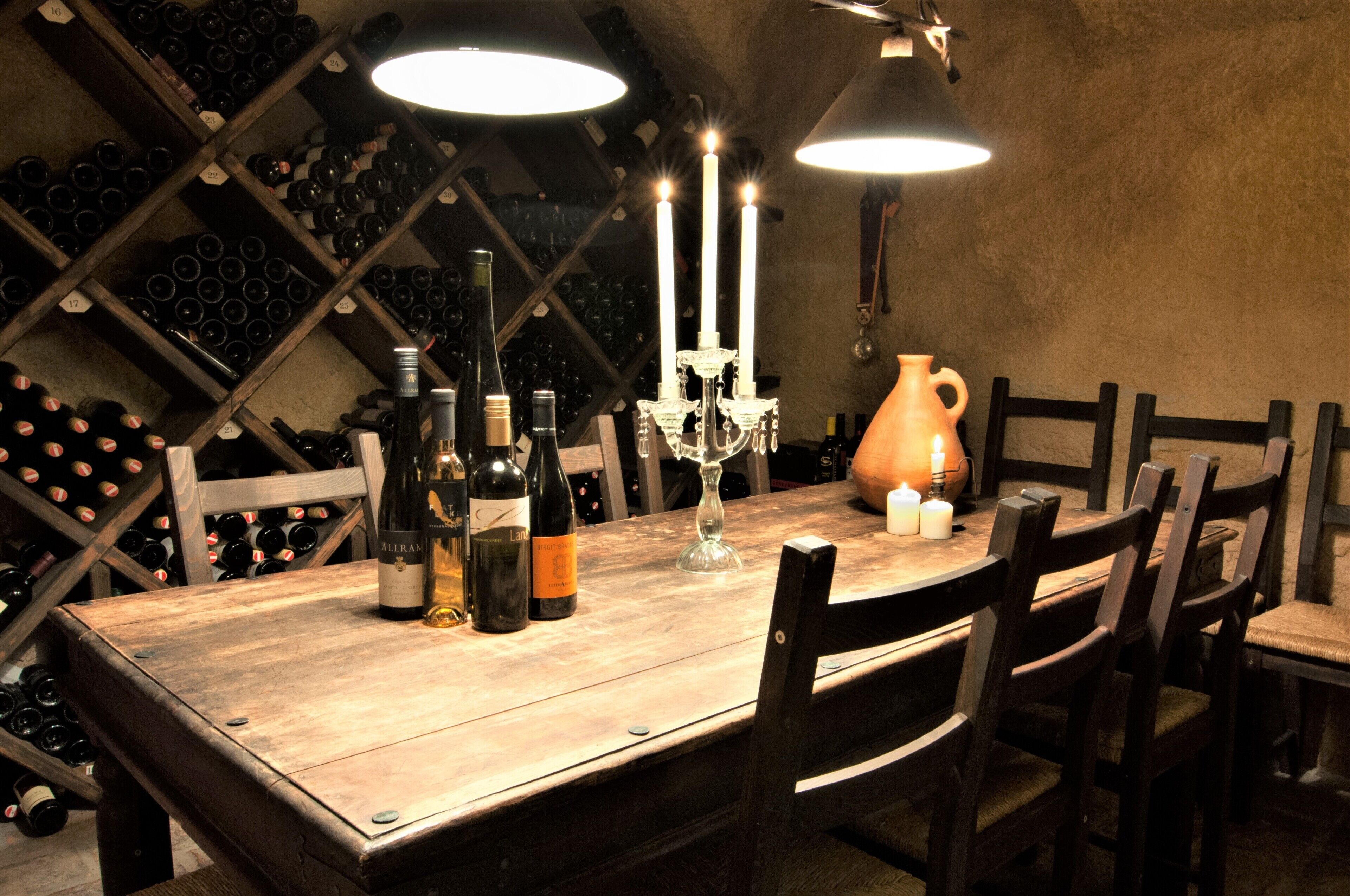 Una mesa de madera con botellas de vino, un candelabro y un florero en una habitación con poca luz y estanterías llenas de vino al fondo.