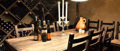Una mesa de madera con botellas de vino, un candelabro y un florero en una habitación con poca luz y estanterías llenas de vino al fondo.