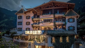Um hotel com vários andares, varandas, uma placa com a inscrição 'Hotel Alpenblick' e montanhas ao fundo.