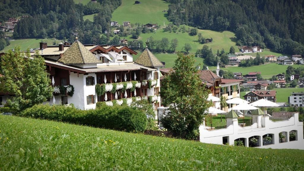 A traditional chalet-style building with a balcony, surrounded by lush greenery and a hilly landscape.