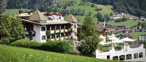 A traditional chalet-style building with a balcony, surrounded by lush greenery and a hilly landscape.