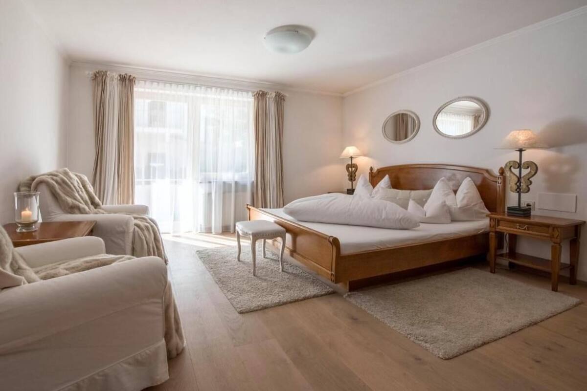 A bedroom with a wooden bed, two bedside tables with lamps, a bedside mirror, a chair, and a window with curtains.