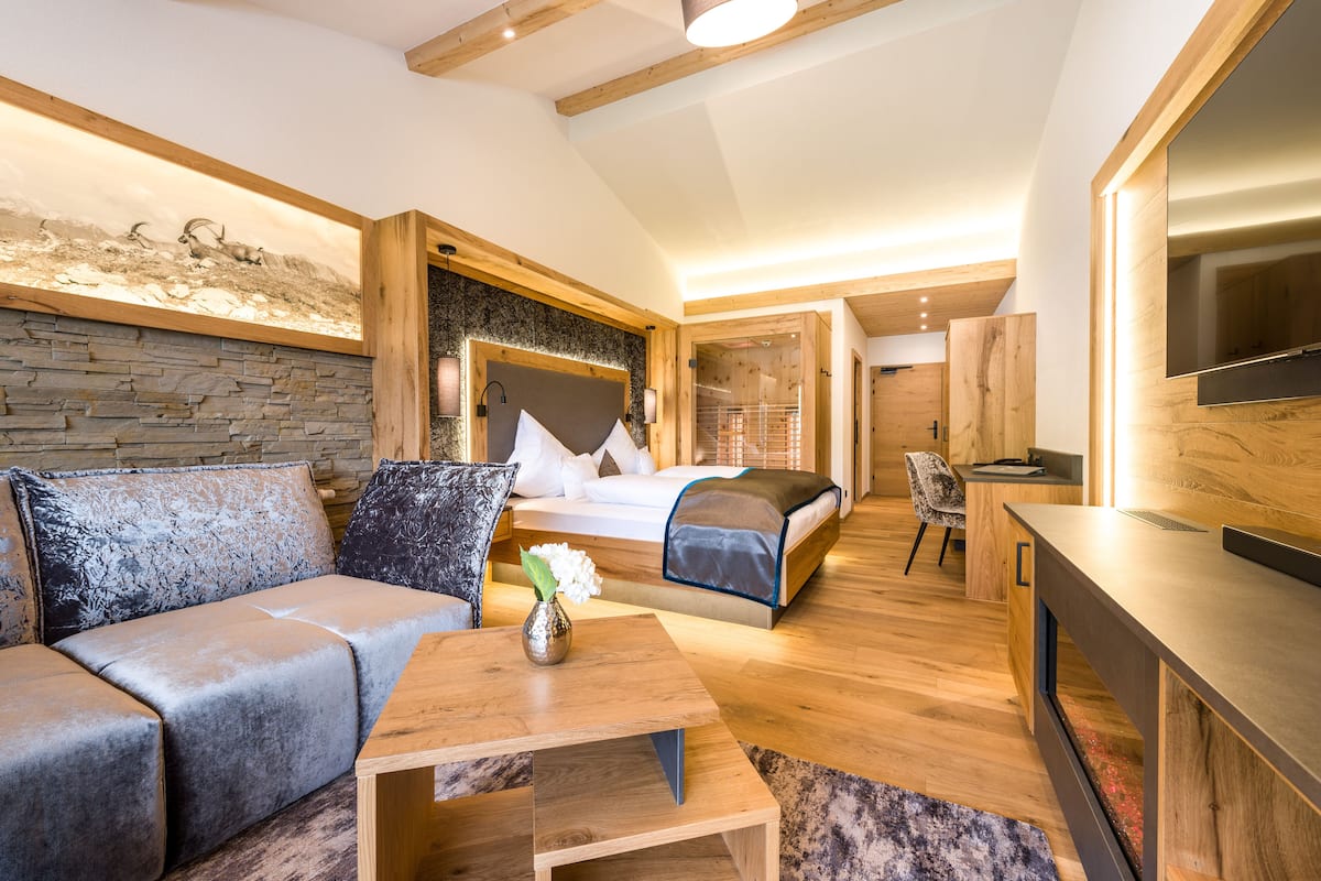 A modern hotel room with a stone wall, a bed with a wooden headboard, a sofa, a small table with a vase, and a TV mounted on the wall.