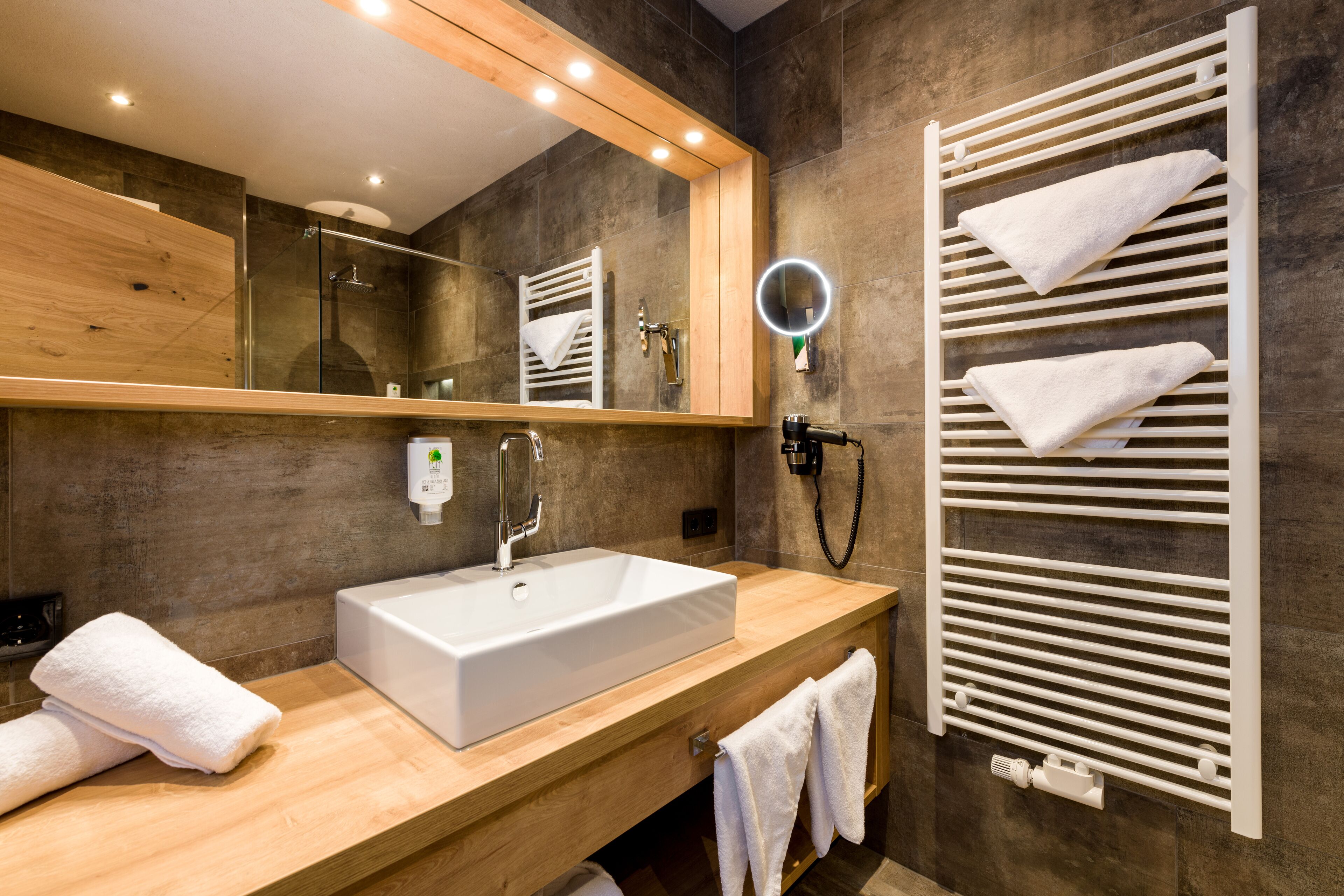 Un baño moderno con tocador de madera, lavabo blanco, cabina de ducha de vidrio y toallero con varias toallas.
