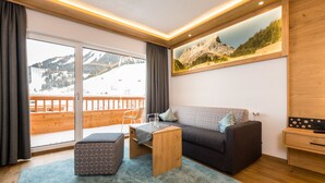 Una sala de estar moderna con un sofá, un reposapiés y una mesa de centro, un ventanal con vistas a una montaña nevada y un cuadro de un paisaje montañoso.