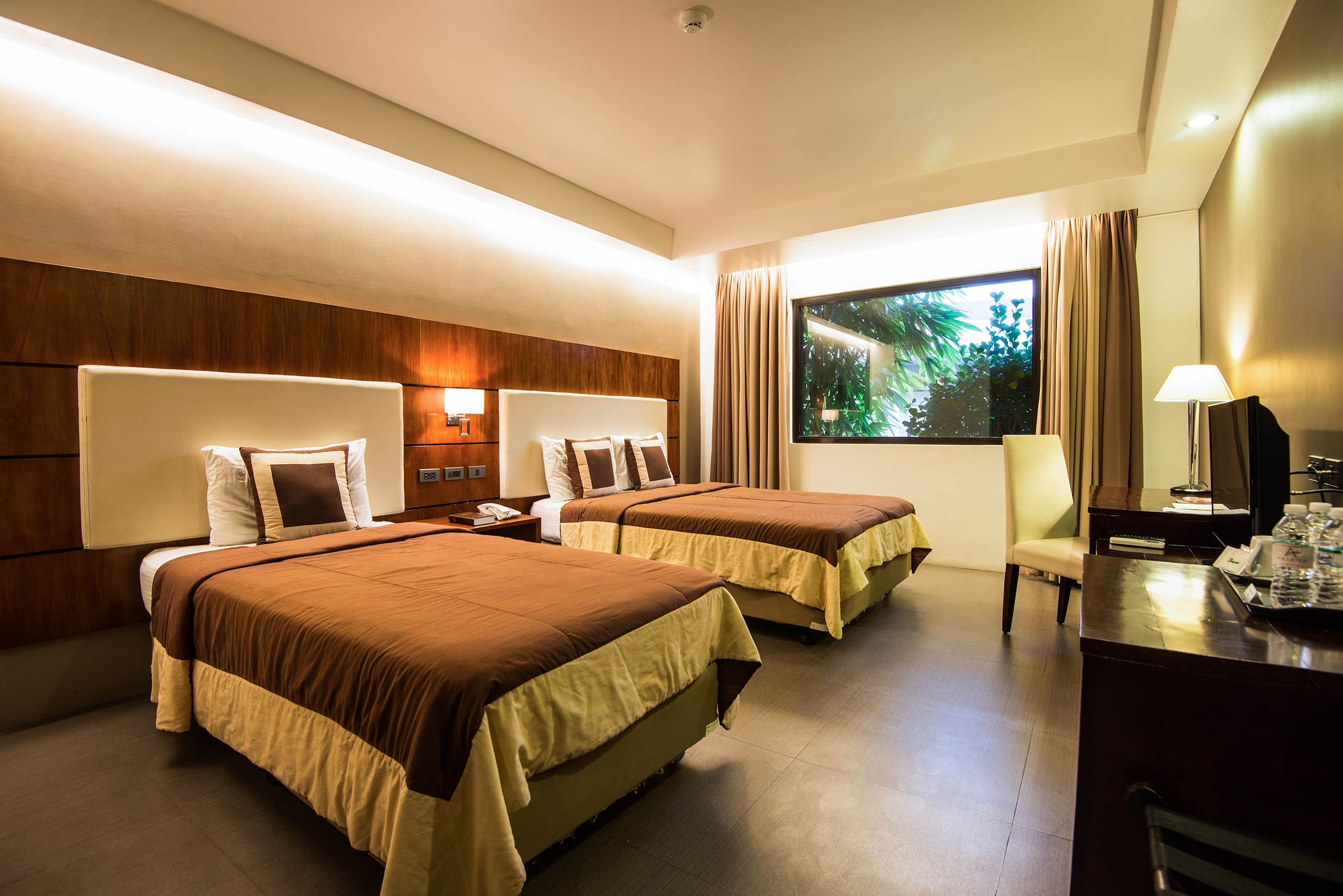 Habitación de hotel con dos camas, un escritorio, una silla, un televisor y una ventana con vistas a zonas verdes.