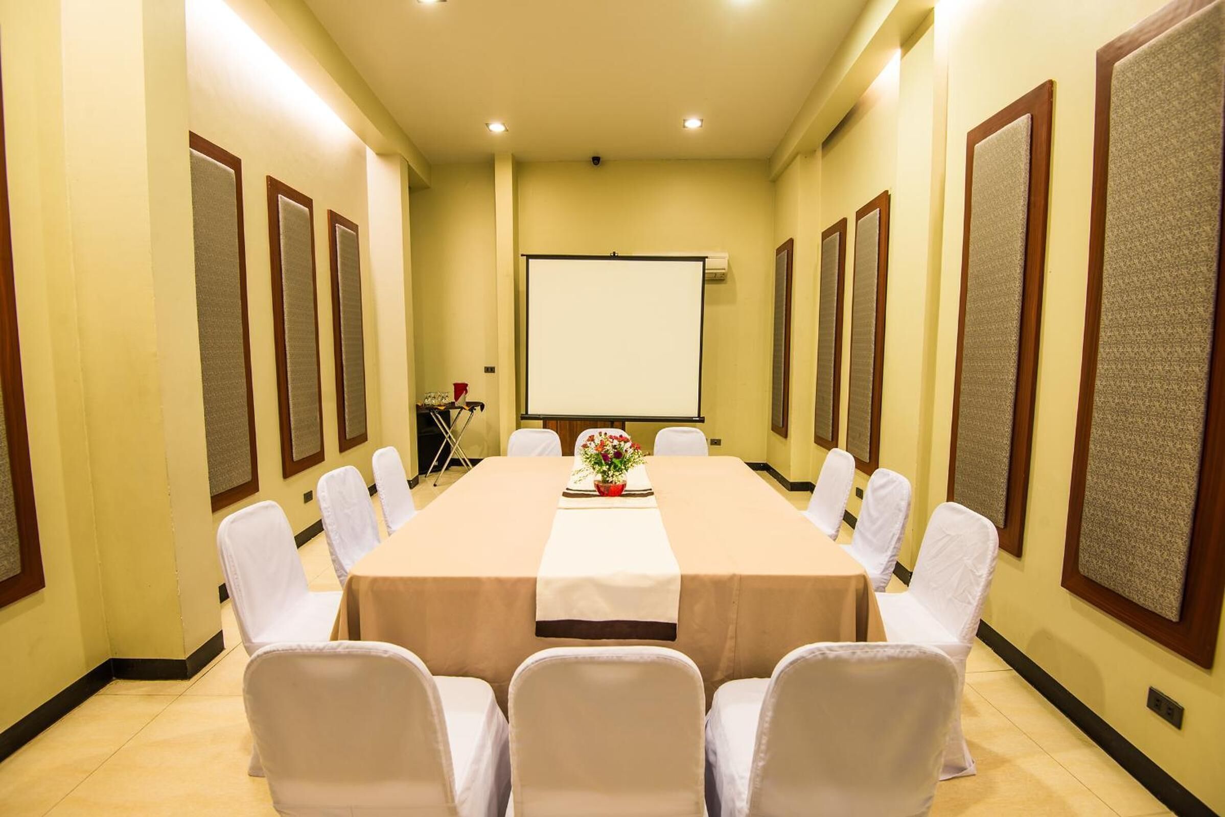 Un salón de conferencias con una larga mesa, sillas blancas y una pantalla para proyector.