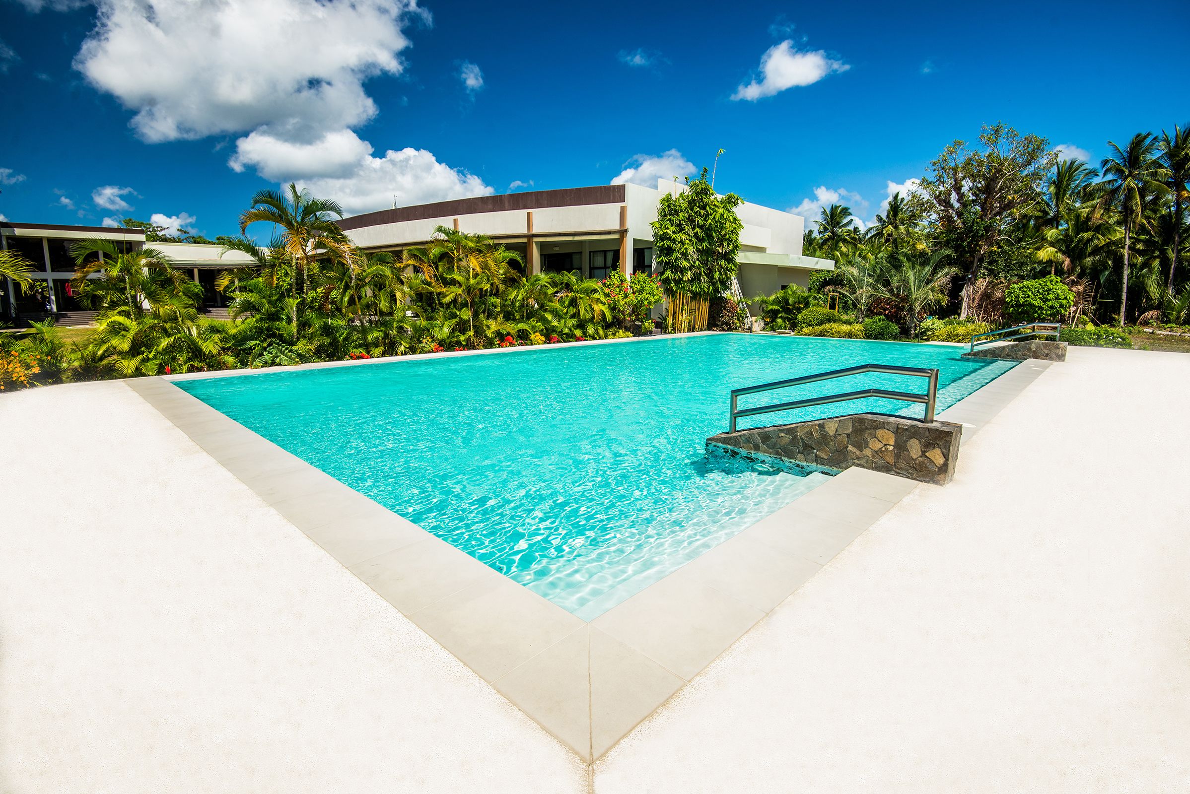 A clear blue pool with a stone border, surrounded by lush greenery and a modern building in the background.