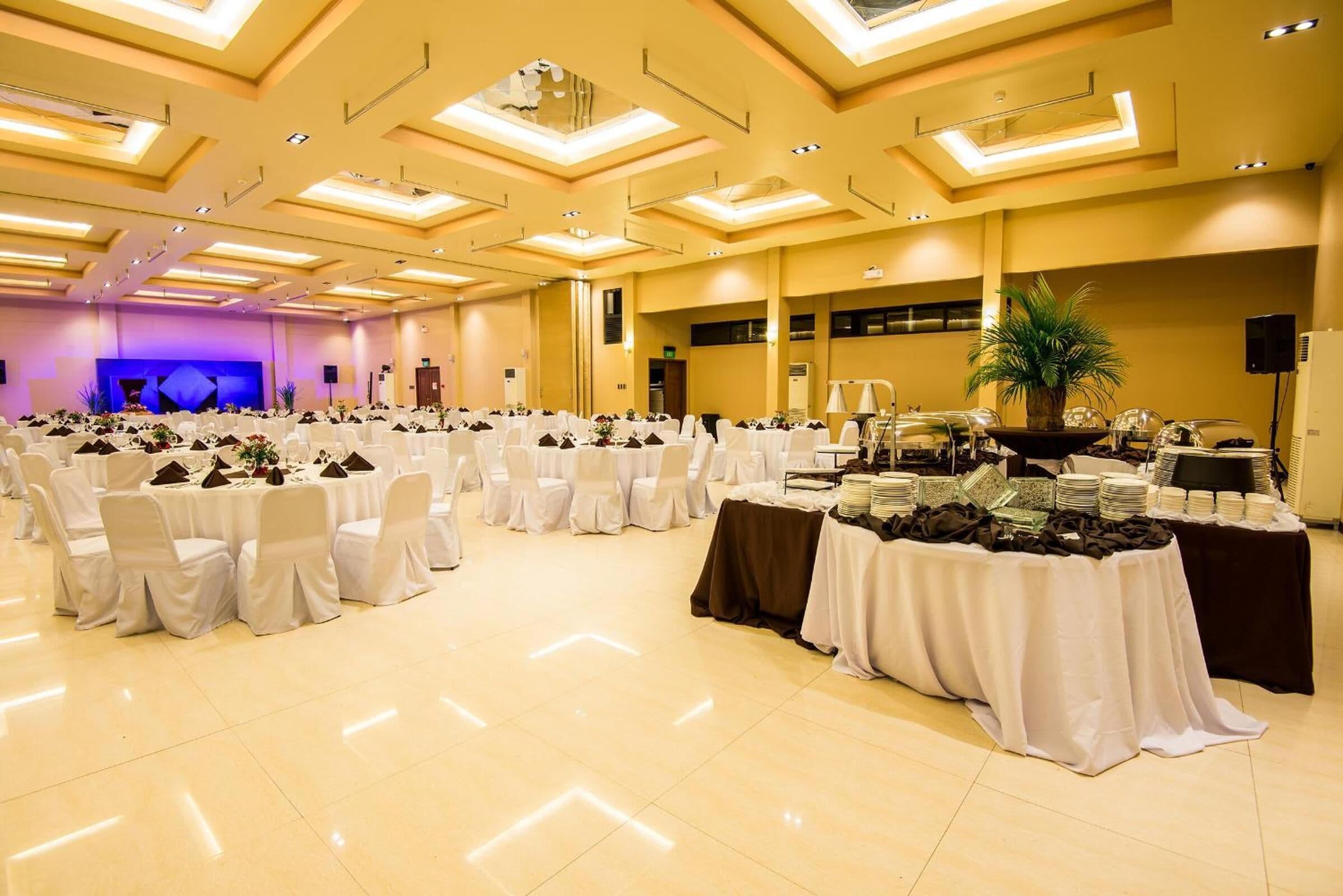 Una vasta sala da banchetti con numerosi tavoli rotondi ricoperti da tovaglie bianche e sedie, un palco con sfondo viola e un'area buffet con diversi piatti.