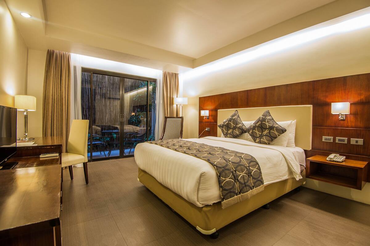 A hotel room with a large bed, a desk, a chair, a nightstand, and a view of the outdoors.