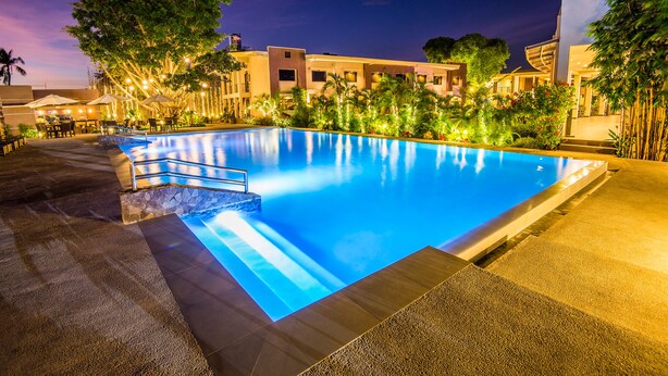 A nighttime swimming pool area with a clear pool, surrounding palm trees, and a building with multiple windows. - Ariana Hotel (Dipolog)