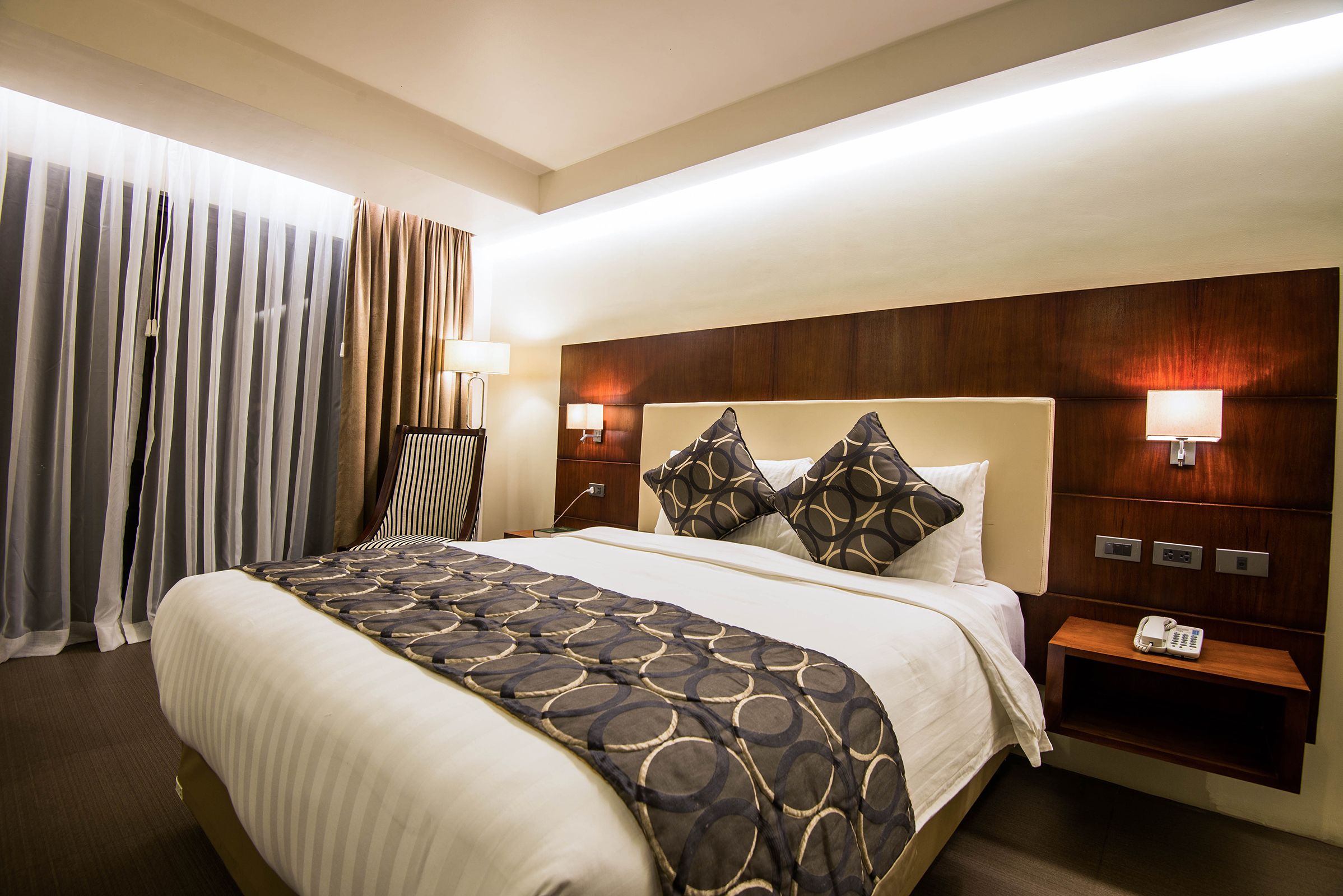 Quarto de hotel com uma cama grande, duas mesas de cabeceira e um criado-mudo com telefone, além de uma cortina na janela.