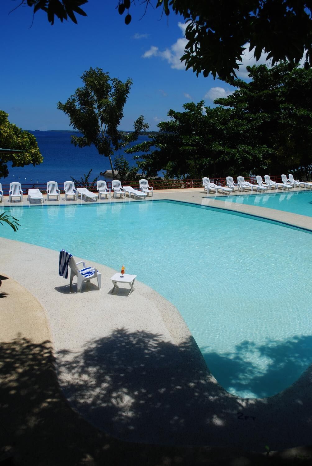 Área junto à piscina com cadeiras de lounge brancas e uma mesa, rodeada por árvores e vista para o mar.
