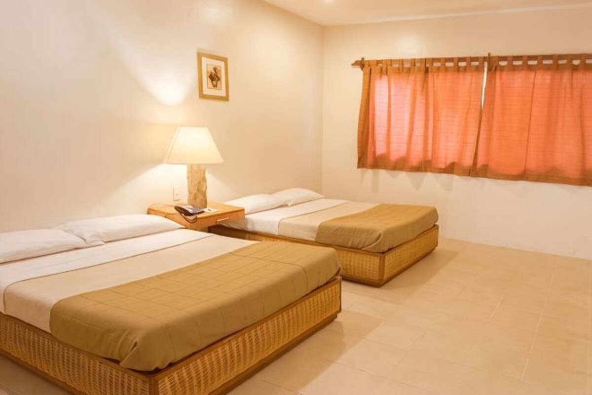 A room with two single beds, a nightstand with a lamp, a window with curtains, and a framed picture on the wall.