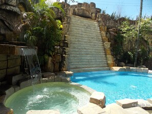 A swimming pool with a waterfall feature and a hot tub area, surrounded by tropical plants and stone walls. - Goldenfield Kundutel Hotel (Bacolod)