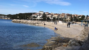 Una zona costera con un paseo marítimo empedrado a lo largo del agua, una playa de arena y edificios al fondo.