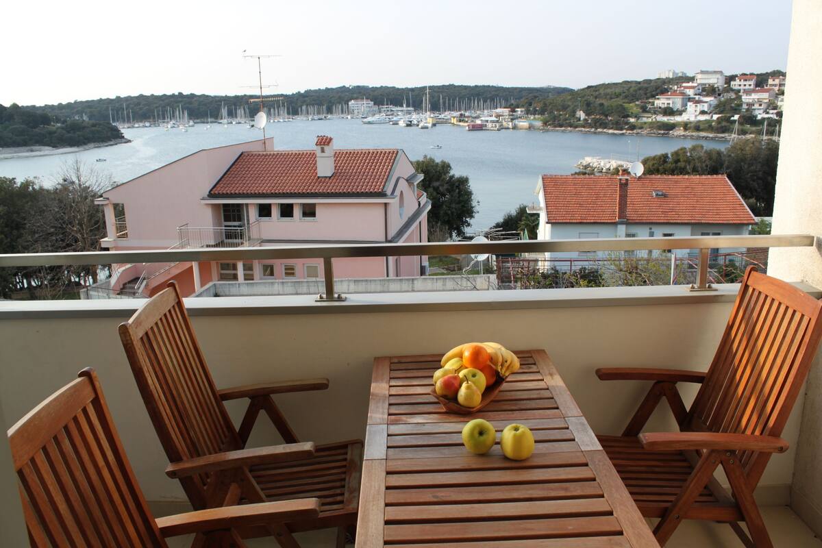 Un balcón con sillas y mesa de madera, vista de un puerto deportivo con barcos y edificios al fondo.