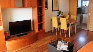 Un espacio habitable compacto con televisor de pantalla plana, armarios de madera, zona de comedor con sillas amarillas y pequeña cocina.