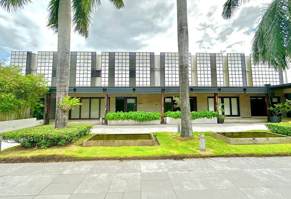 Un edificio moderno con fachada simétrica, ventanas amplias y un jardín cuidado con palmeras y una fuente de agua.