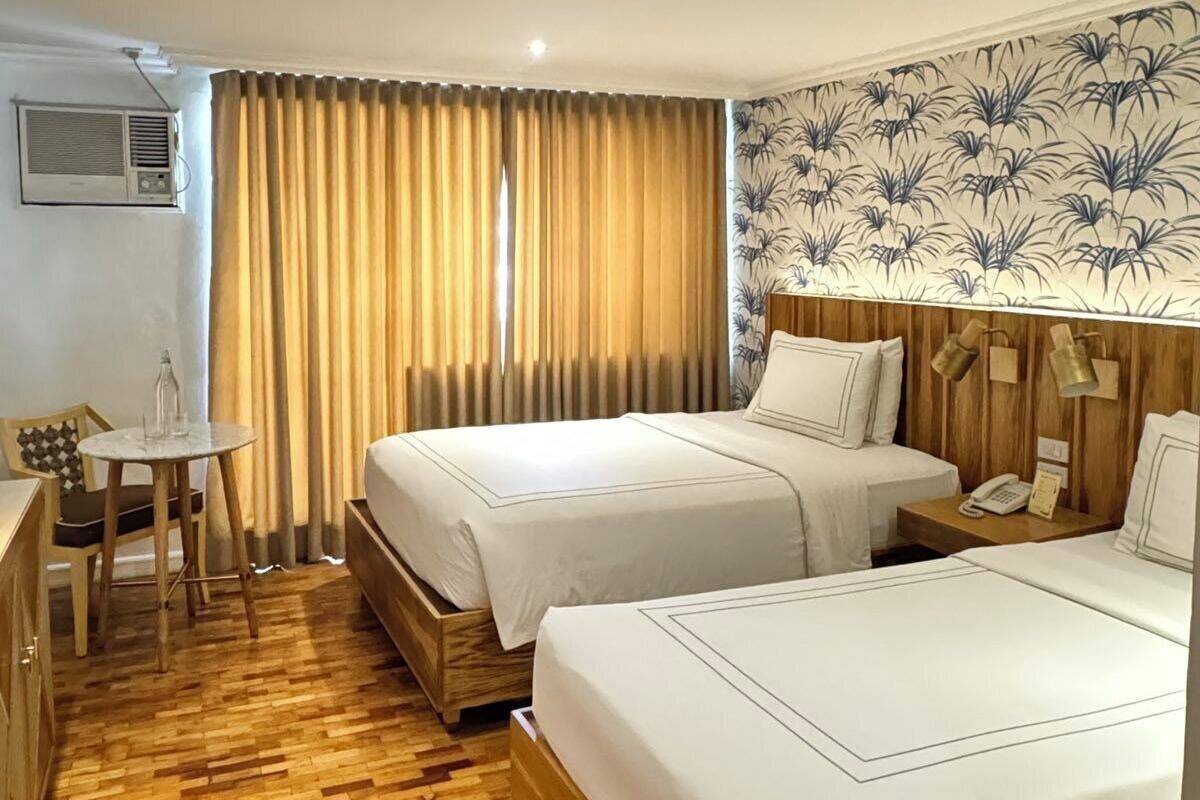 Une chambre d’hôtel avec deux lits, un plancher en bois, une petite table avec une chaise, un climatiseur fixé au mur et un papier peint à motifs.