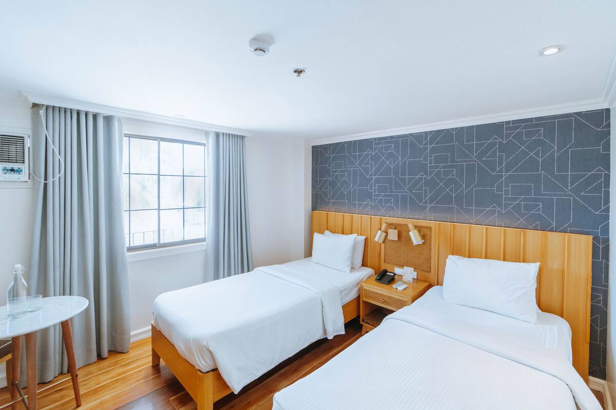 Une chambre d’hôtel avec deux lits, une petite table, une fenêtre avec des rideaux et un mur orné d’un motif géométrique.