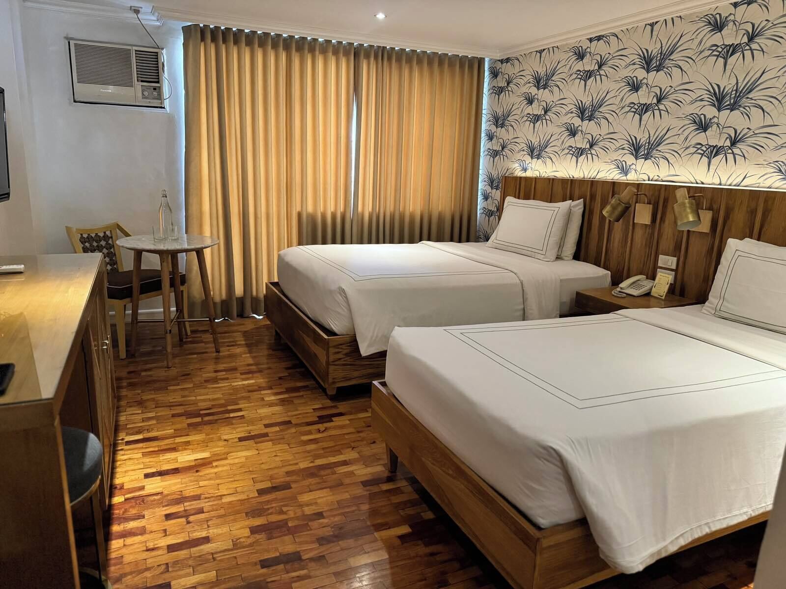 A hotel room with two beds, a wooden floor, a small table with chairs, and a curtain.