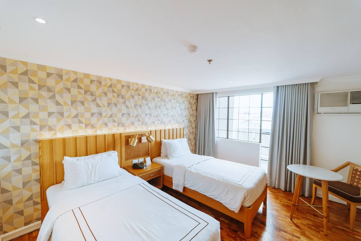 Une chambre d’hôtel avec deux lits, une tête de lit en bois, une petite table et une fenêtre avec des rideaux.