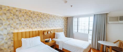 Habitación de hotel con dos camas, cabecero de madera, una mesita y una ventana con cortinas.