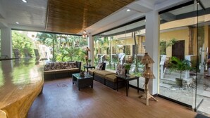 A spacious living area with a wooden bench, a sofa, and a coffee table. There are large windows offering a view of greenery outside. - Bamboo Beach Resort and Restaurant (Balabag)