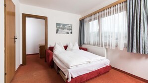 Une chambre d’hôtel avec un lit, un canapé rouge, une porte en bois, une fenêtre avec des rideaux et un tableau accroché au mur.