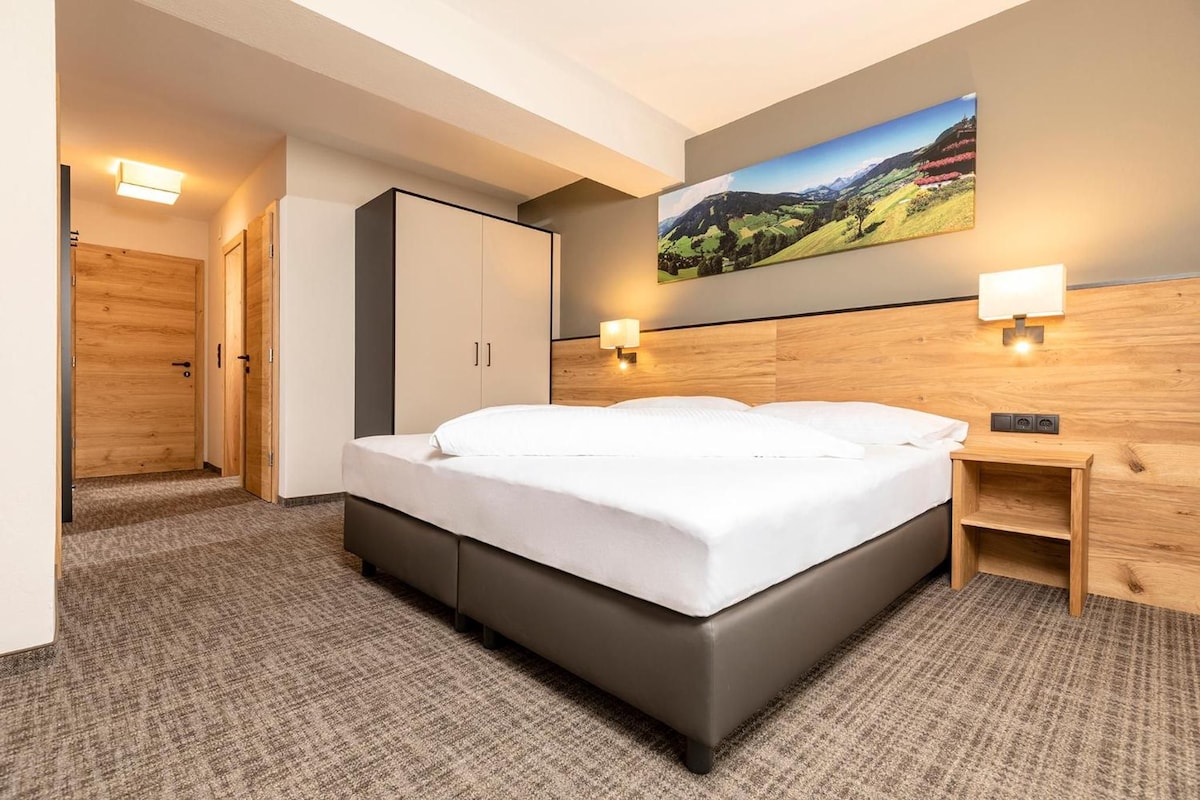 Une chambre d’hôtel avec un lit, une table de chevet, une lampe fixée au mur et un tableau représentant un paysage.