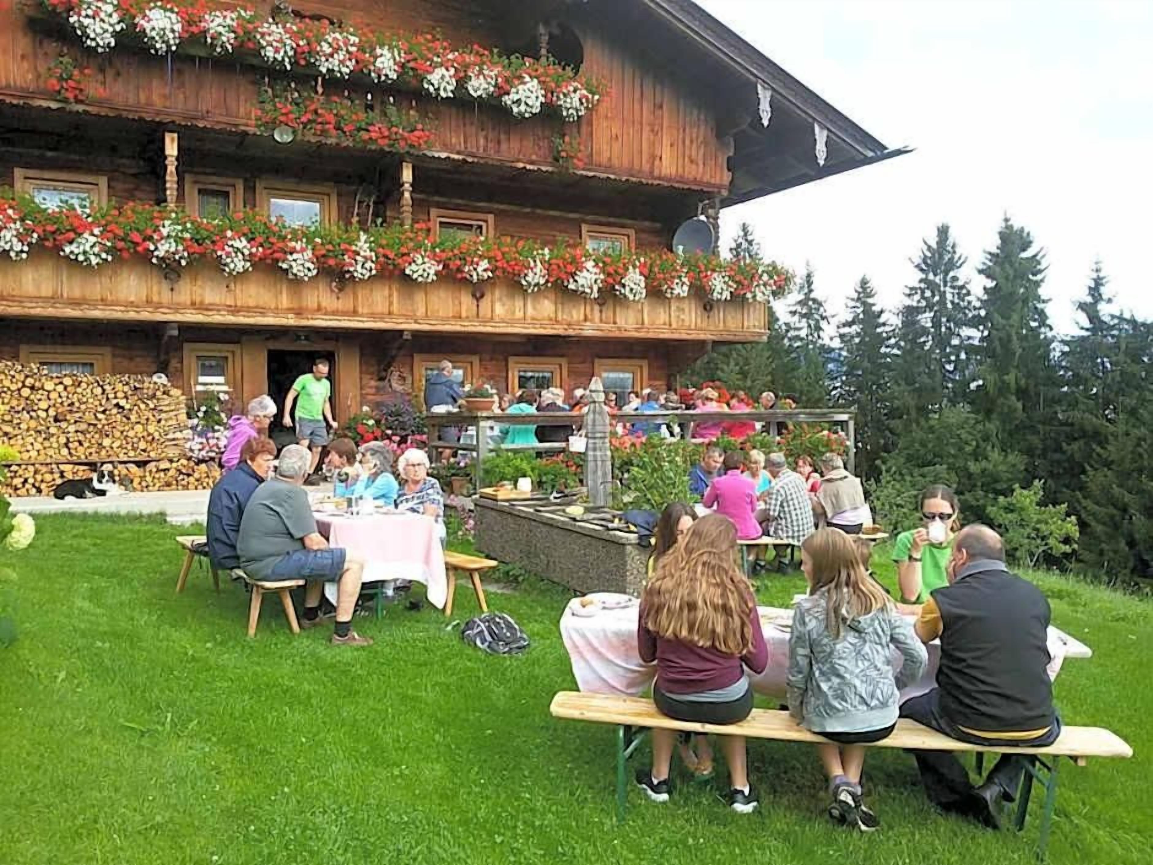 Un área de comedor al aire libre con mesas y sillas, un edificio de madera con jardineras de flores, y una persona de pie en el balcón.