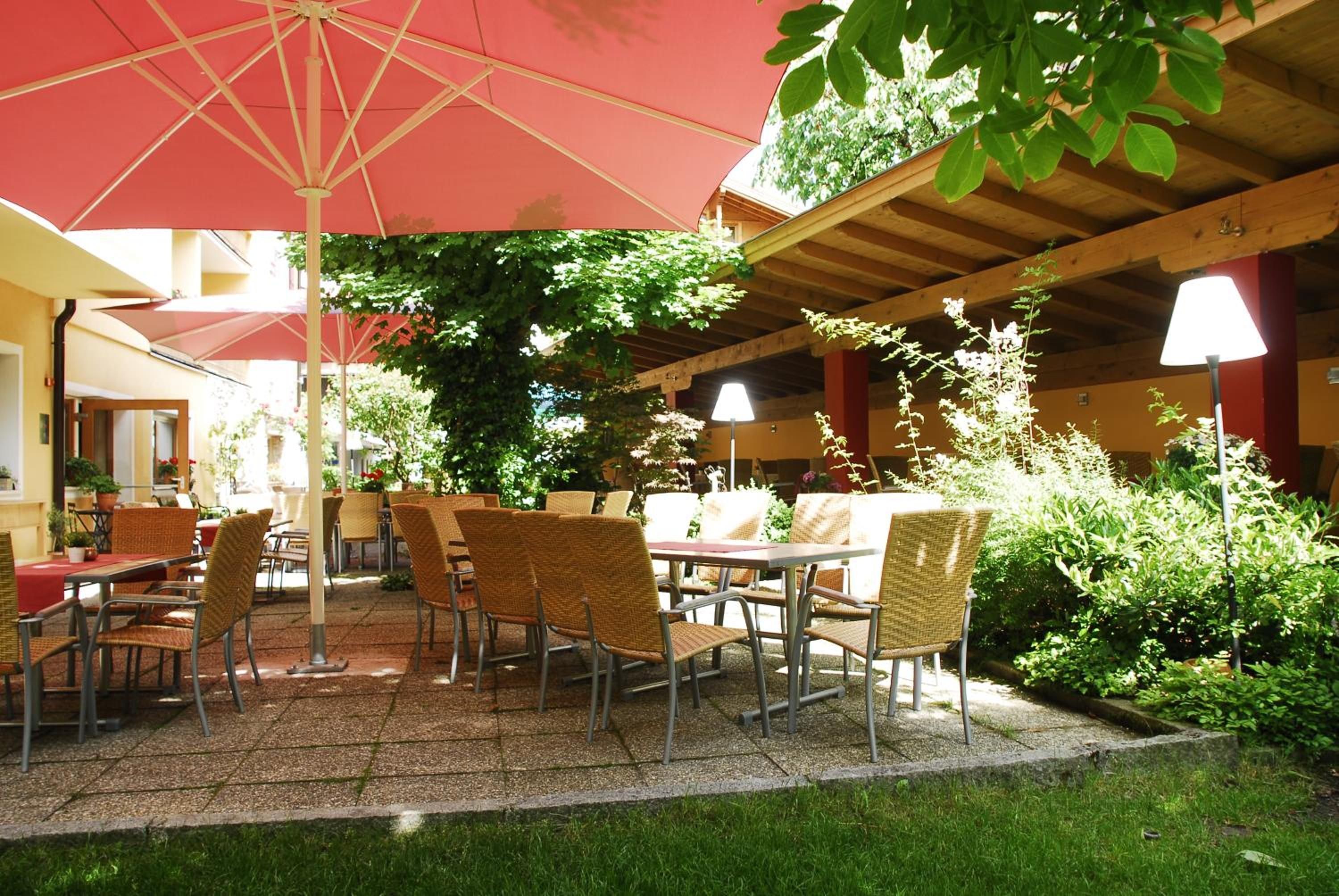 En uteplats med bord och stolar, skyddad av ett stort parasoll, omgiven av grönska och en träpergola.
