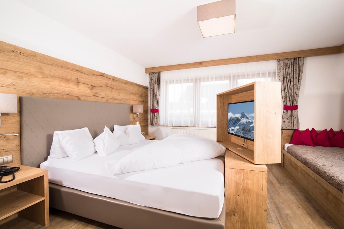 A hotel room with a bed, a TV, a bedside table, and a window with curtains.