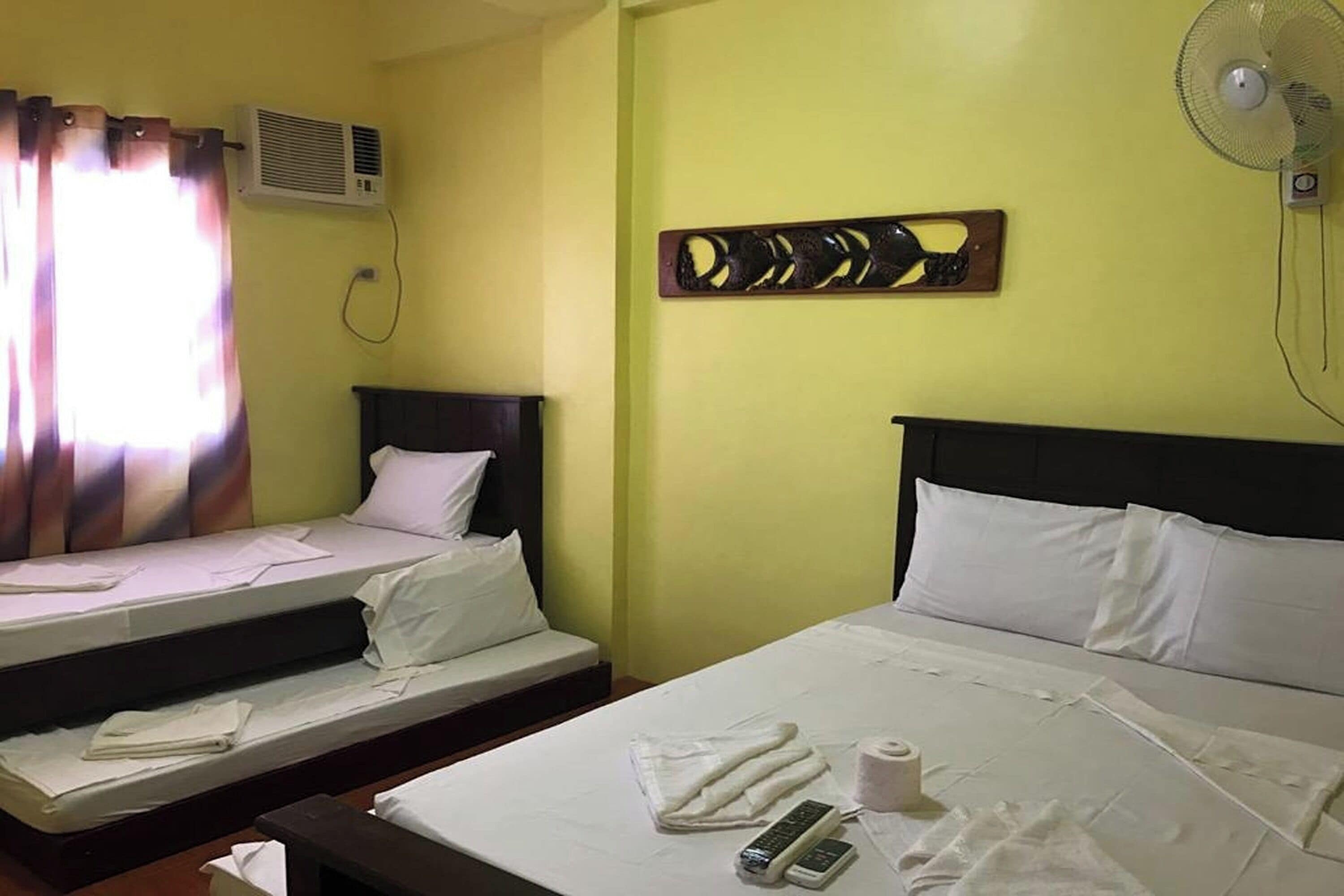A hotel room with two single beds, a large bed, a wall-mounted air conditioner, a ceiling fan, and a decorative wall piece.