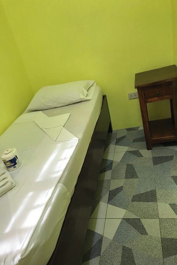 A small, single-bed room with a plain mattress, a wooden nightstand, and a patterned floor.