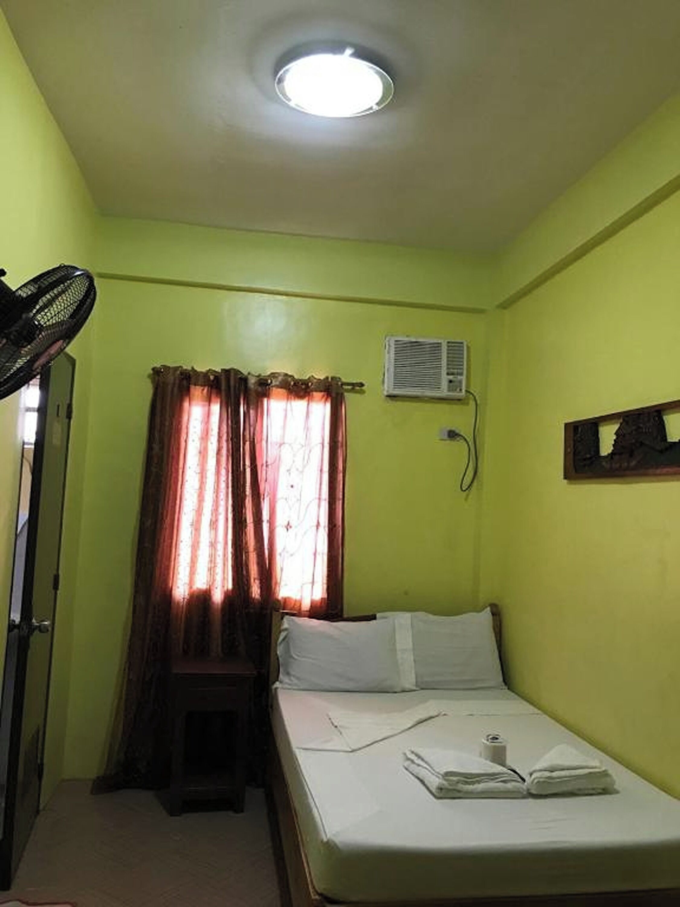 A small, single-bed room with a ceiling fan, an air conditioning unit, and a window with curtains.