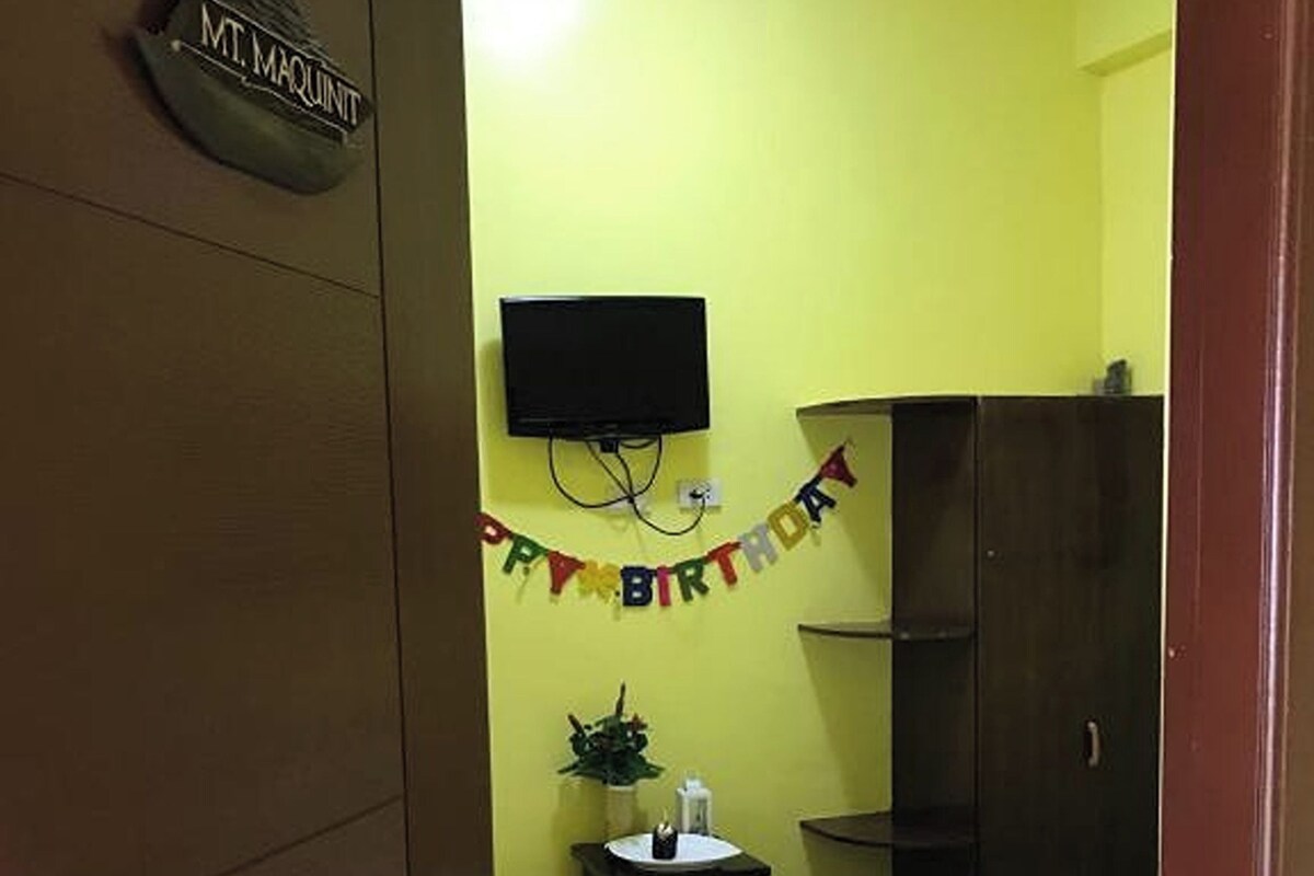 Quarto com uma cama, televisão fixada na parede, uma mesinha com uma planta e um banner de 'Feliz Aniversário'.