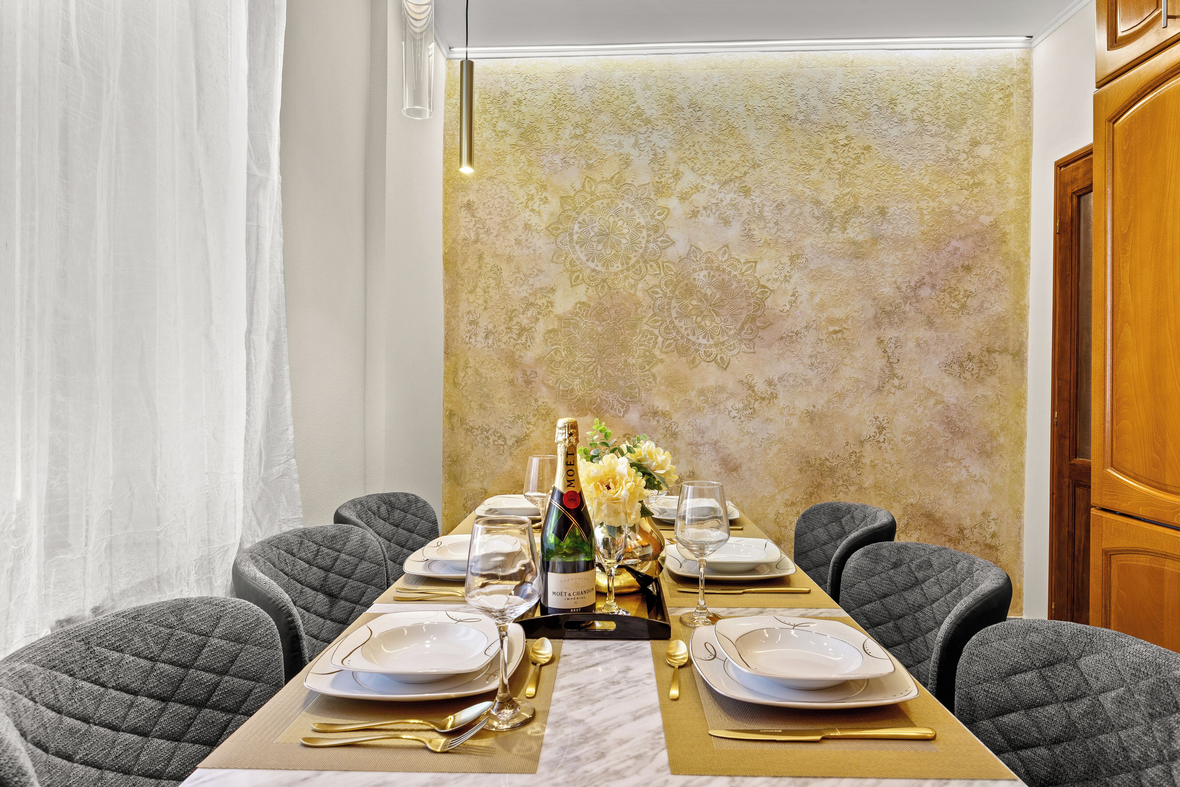 Une table à manger dressée pour six personnes, avec une bouteille de champagne, des couverts dorés et un centre de table floral.