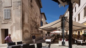 An outdoor café with tables and chairs, a stone building, and a clear blue sky. - Diana City Hall View (Pula)