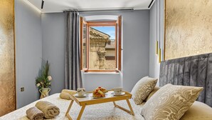 A neatly made bed with a tray of breakfast, a window with a view of a building, and a decorative vase with flowers. - Diana City Hall View (Pula)