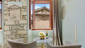 A room with a stone wall, a window with a view of a building, a round table with a bottle and flowers, and two chairs. - Diana City Hall View (Pula)